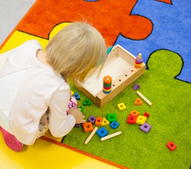 Na kolorowym dywanie tyłem kuca dziecko. Bawi sie drewnianymi, kolorowymi klockami