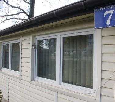 Na zewnątrz, dzien. Na niskim domu z białego sajdingu w oknie zaczepiony sensor
