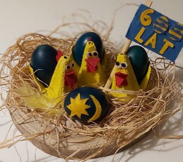 Trzy kurki z granatowymi jajkami w gnieździe, jedna z nich z flagą 600-lecia. Przed nimi pisanka z herbem miasta.