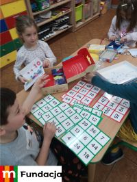 Dzieci siedzą przy stolikach, z pudełek wyjmują łamigłówki matematyczne, logo MFundacji w lewym dolnym rogu