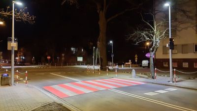 Na zewnątrz, noc. Przejście dla pieszych biało-czerwone oświetlone po obu stronach latarniami. Po prawej blok