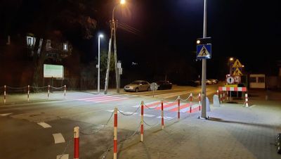Na zewnątrz, noc. Przejście dla pieszych biało-czerwone oświetlone po obu stronach latarniami. Po prawej znaki z robotami...