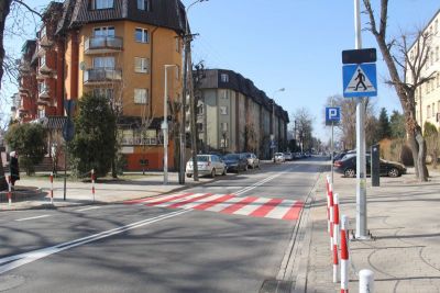 Na zewnątrz, dzień. Biało-czerwone przejście dla pieszych na ulicy. Po obu stronach chodniki i budynki mieszkalne