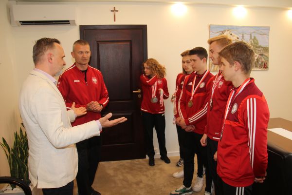 Oświetlony gabinet. Sześciu zawodników w czerwonych bluzach. Jeden mężczyzna w jasnej marynarce. Rozmawiają