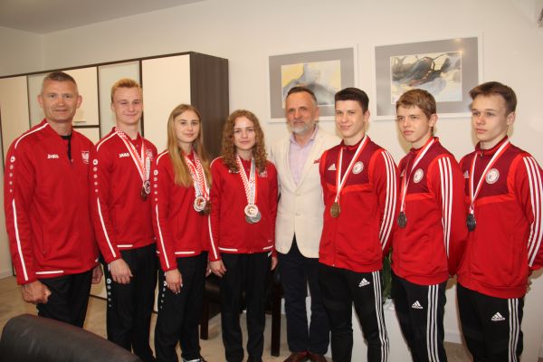 Oświetlony gabinet. Sześciu zawodników w czerwonych bluzach. Jeden mężczyzna w jasnej marynarce