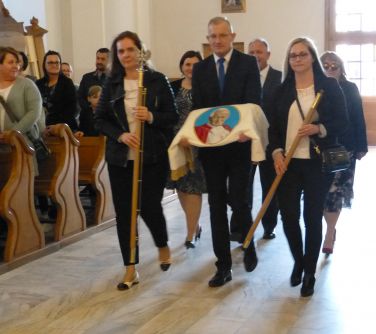 Wnętrze kościoła. Środkiem maszerują trzy osoby. Mężczyzna w środku niesie złożony sztandar.