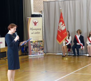 Jasna aula. Kobieta przemawia do mikrofonu. Po prawej na krzesłach siedzą dzieci. Ubrani są na galowo