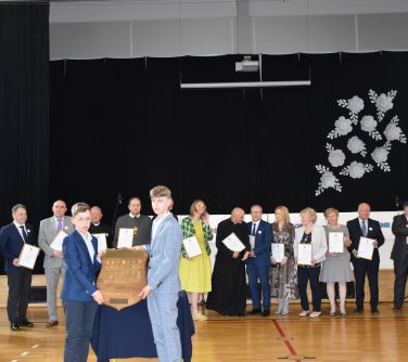Jasna aula. Grupa osób. W środku dwóch chłopców w garniturach. trzymają herb