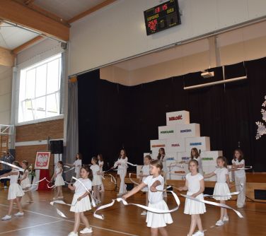 Jasna aula. Dzieci ubrane na biało machają szarfami