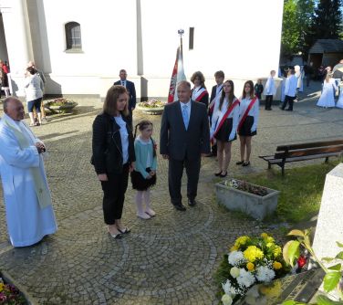 Na dworze, przed kościołem. Słońce. Przed krzyżem ksiądz, kobieta, dziecko i mężczyzna