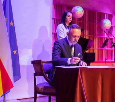 Mężczyzna na scenie przy stoliku podpisuje dokumenty