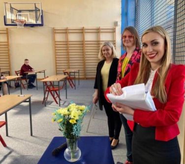 Hala sportowa. Trzy kobiety. Jedna w czerwonej marynarce trzyma arkusze