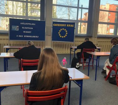 Aula szkolna. Widok na okna. Dzień. W ławkach szkolnych tyłem siedza osoby