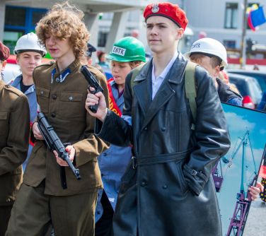 Dzień, słońce. Po ulicy maszerują osoby w pzebraniach. Na przodzie jeden żołnierz, drugi gankster