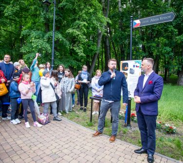 Alejka w parku za dnia. Po lewej grupa osób zgromadzona. Na przodzie przemawia mężczyzna