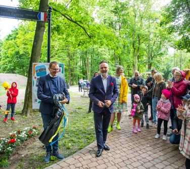 Alejka w parku za dnia. Po prawej grupa osób. na przodzie dwóch mężczyzn