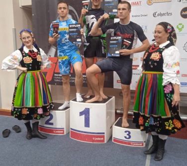 trzech zawodników MMA stoi na podium w towarzystwie dziewcząt w strojach ludowych