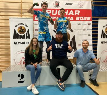 dwóch zawodników stoi na podium, trzy osoby siedzą na pierwszym planie