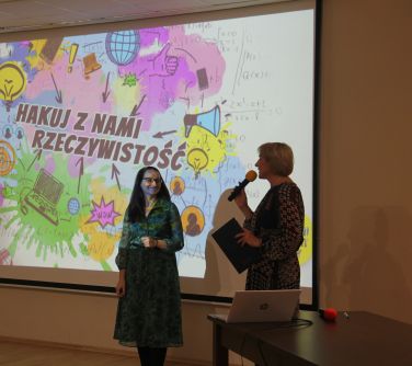 kobieta w towarzystwie drugiej kobiety przemawia do mikrofonu