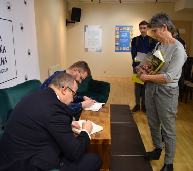mężczyźni podpisują przy stoliku książki po prawej stronie stoi kobieta i oczekuje na autograf
