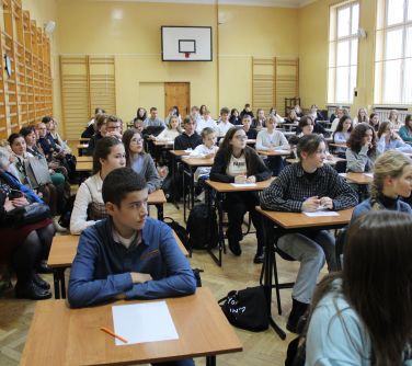 dzieci siedzą w pojedyńczych ławkach na sali gimnastycznej, na stolikach kartki, z lewej strony na ławeczkach rodzice