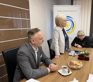 p. Burmistrz siedzi przy stole, po lewej stronie stoi kobieta i przemawia, druga kobieta siedzi, w tle baner miasta