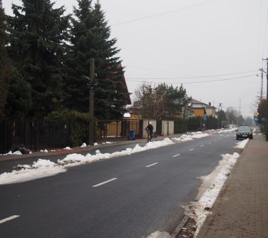 Na zewnątrz pochmurny dzień. Ulica, śnieg po obu stronach krawężników, po prawej samochód, po lewej po chodniku jedzie osoba
