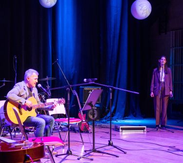 na scenie po prawej stronie na krześle siedzi mężczyzna i gra na gitarze, po lewej stronie stoja dwie nastolatki i śpiewają...