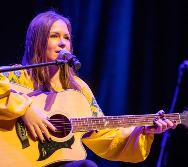 na scenie na krześle siedzi kobieta w żółtej bluzce, trzyma gitarę, gra na niej i śpiewa