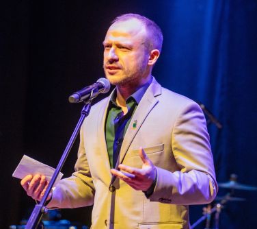Mężczyzna stoi przed mikrofonem, w prawym ręku trzyma kartkę