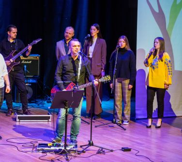 Kobiety i mężczyźni stoją na scenie, trzymają mikrofony, śpiewają, na środku oraz z tyłu mężczyzni grają na gitarach