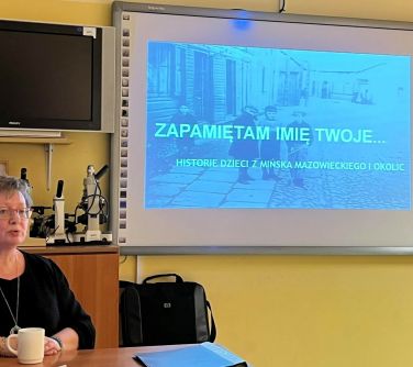 kobieta siedzi na krześle przy biurku po lewej stronie, w tle na projektorze wyświetlany tekst zapamiętam imie swoje