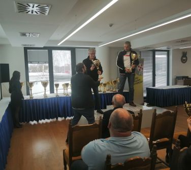 osoby siedzące, dwie stoją na podium trzymają puchary, w tyle na stole przykrytym bialym obrusem,