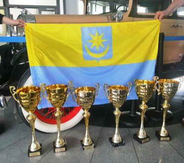 flaga mińska mazowieckiego żółto niebieska trzymana przez osoby, przed flaga stoi 6 pucharów