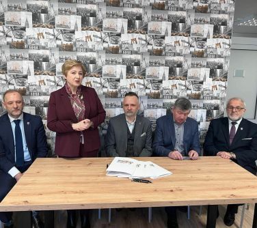 pięć osób przy stole , czterech mężczyzn siedzi, jedna kobieta stoi trzyma ręcę złożóne przemaiwa, na stole leżą dokumenty...