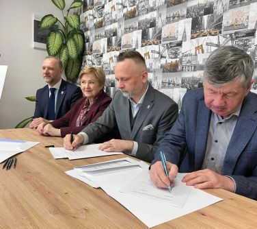 cztery osoby siedzą przy stole trzech mężczyzn i jedna kobieta. Dwóch mężczyzn podpisuje dokumenty