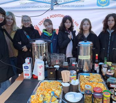 osoby stoją pod namiotem na pikniku, przed nimi stoi stolik z dwoma warnikami, owoce kawa, hebrata mleko cukier