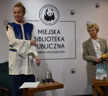 dwie kobiety stoja przed fotelami przy kobietach małe stoliki, na jednym stoi woda w dzbanku i szklanka, na drugim książka...