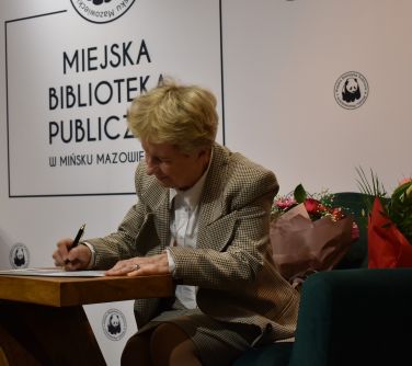 kobieta siedzi na fotelu, podpisuje książkę przy stoliku
