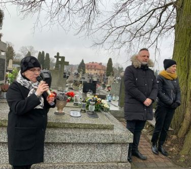 po lewej stronie kobieta przemawia do mikrofonu, po prawej stronie mężczyzna stoi z kobietą między grobami na cmentarzu