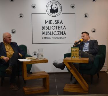 dwóch mężczyzn siedzi na scenie na fotelach jeden przemawia do mikrofonu