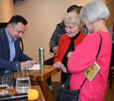 mężczyzna podpisuje ksiązkę przed nim stoją kobiety