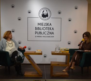 dwie kobiety siedzą naprzeciwko siebie w fotelach, jedna z nich przemawia do mikrofonu