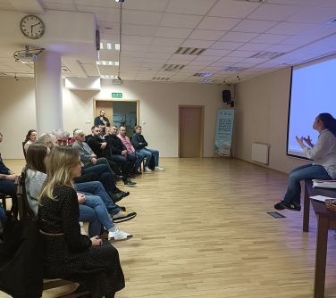 osoby siedzą w półkolu, kobieta siedzi przed nimi na stole
