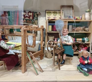 miniaturka kobiety siedzi przy małym kole przędzie, naokoło miniaturowe meble