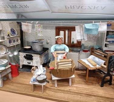 miniaturowa kobieta pierze na miniaturowej tarze, naokoło miniaturowe meble i garnki, patelnie, misy