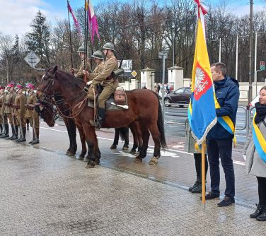trzy osoby stoją ze sztandarem, obok kawaleria na koniach