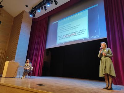 dwie kobiety na scenie po lewej stronie kobieta siedzi na krześle przed mównicą, po prawej stoi i przemawia do mikrofonu