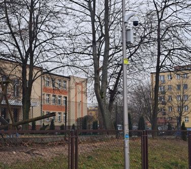 Na zewnątrz, pochmurny dzień. Zdjęcie latarni z przyczepionym monitoringiem, w tle szkoła