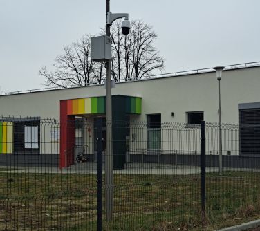 Na zewnątrz, pochmurny dzień. Zdjęcie latarni przy ogrodzeniu z zamontowanym monitoringiem, w tle przedszkole